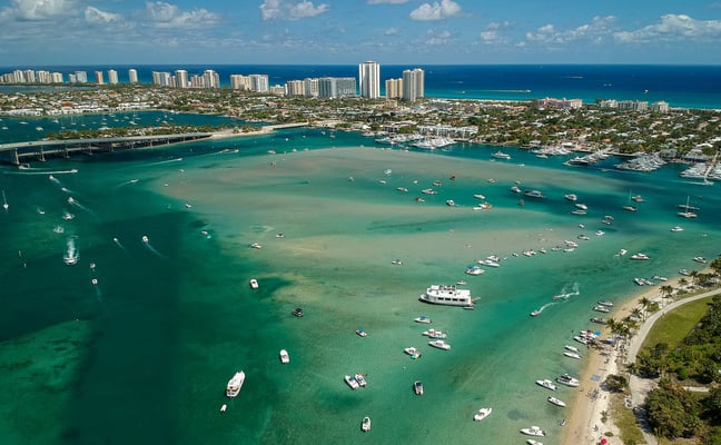 Aerial view of turquoise inlet with moored boats, sandy beach, bridge, and high-rise buildings along the shoreline